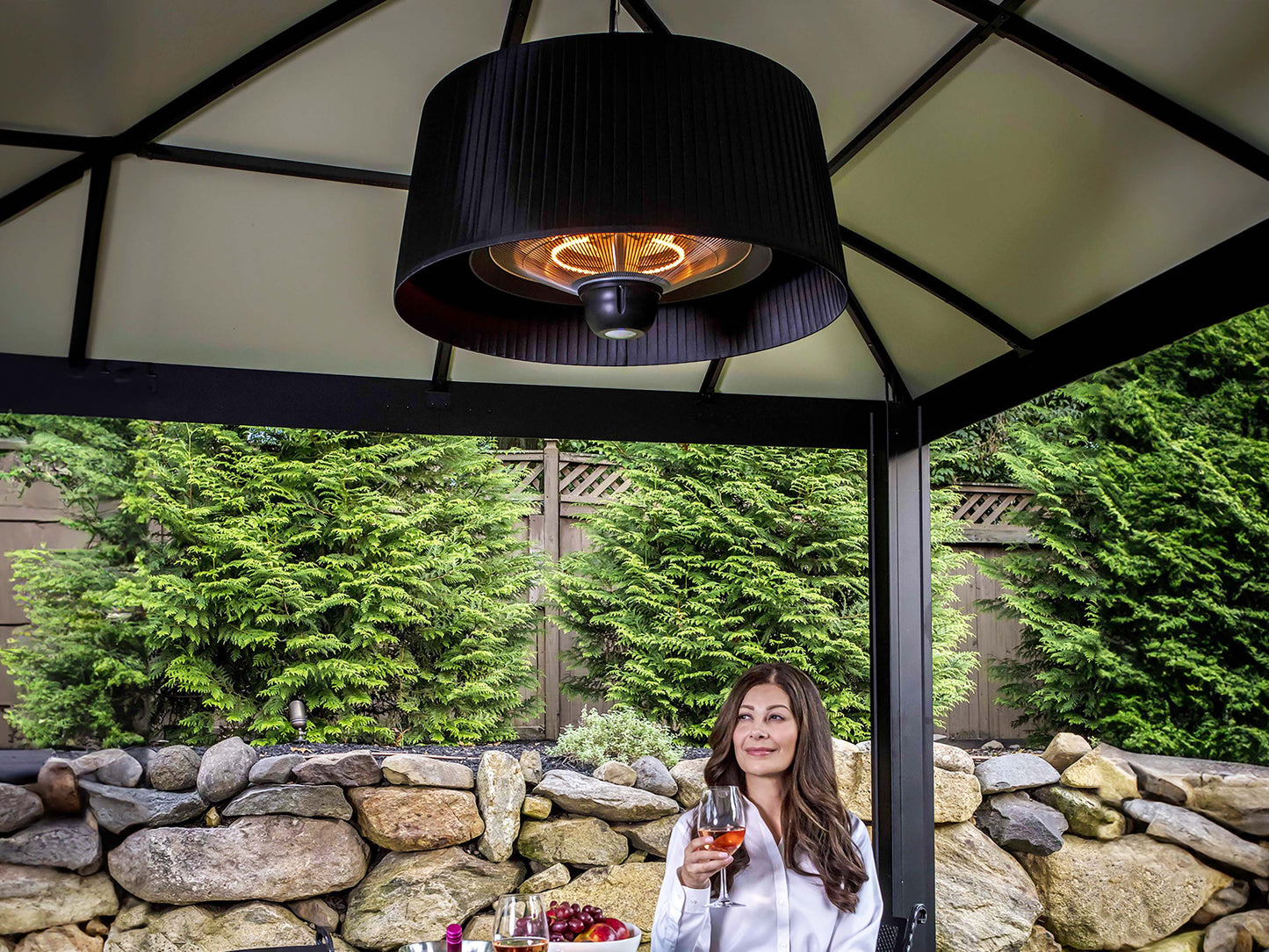 Glow Pendent Heat Lamp hanging from gazebo. Woman sitting underneath with glass of wine.